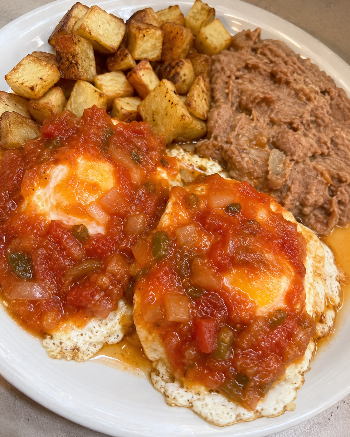 Huevos Rancheros with onions, tomatoes, and jalapeño peppers, served on a white plate with a side of refried beans and potatoes