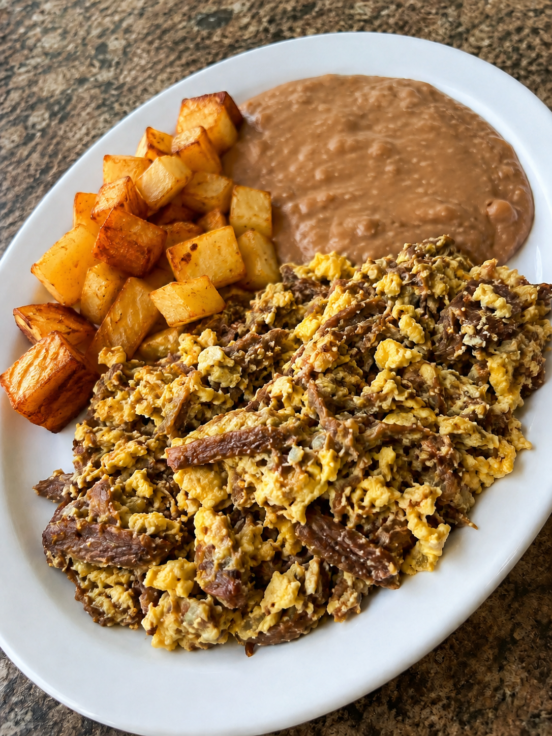 Machacado con Huevo with shredded beef, scrambled eggs, onions, tomatoes, and jalapeño peppers, served on a white plate with a side of refried beans and potatoes