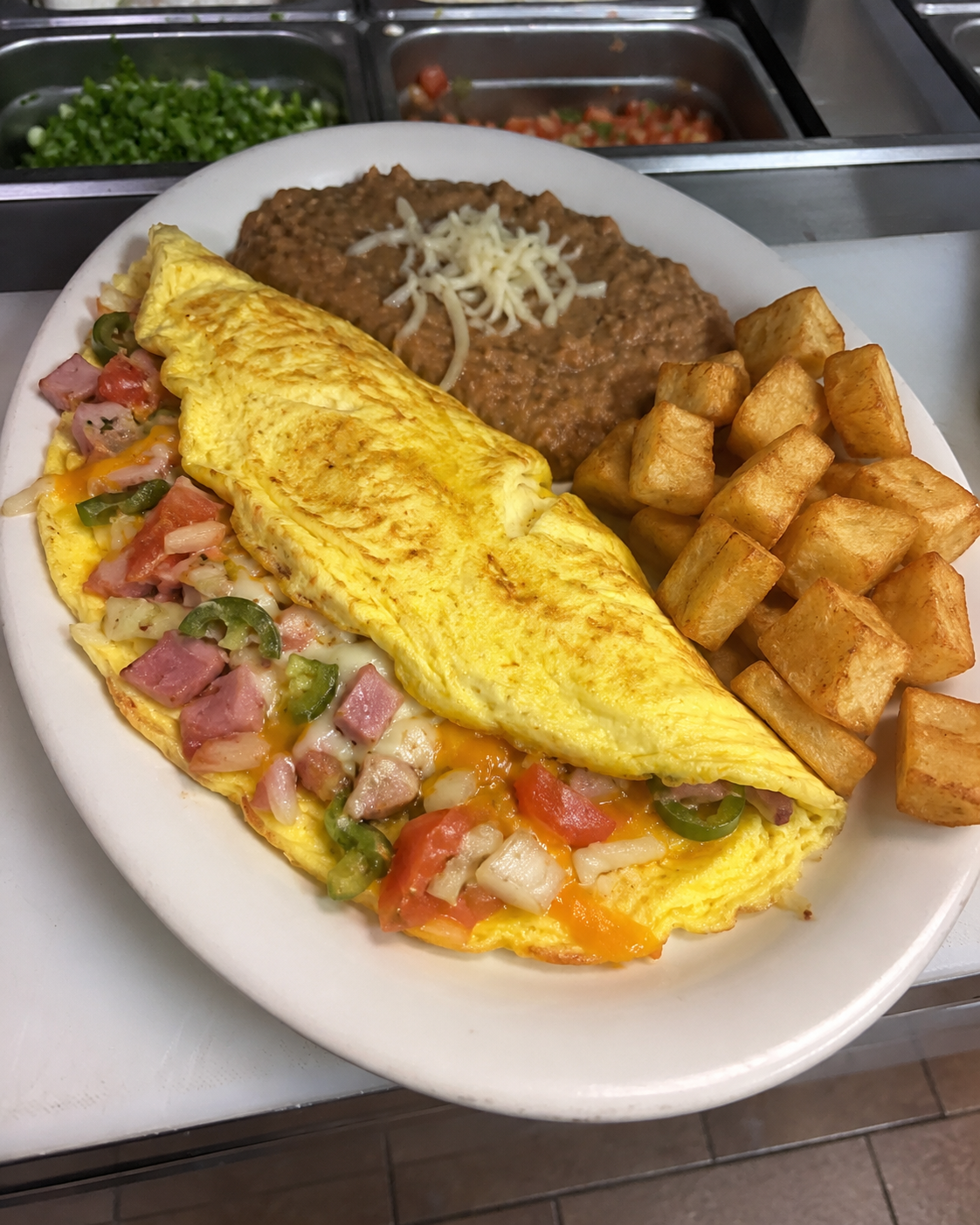 Omelette with onions, tomatoes, and jalapeño peppers, served on a white plate with a side of refried beans and potatoes