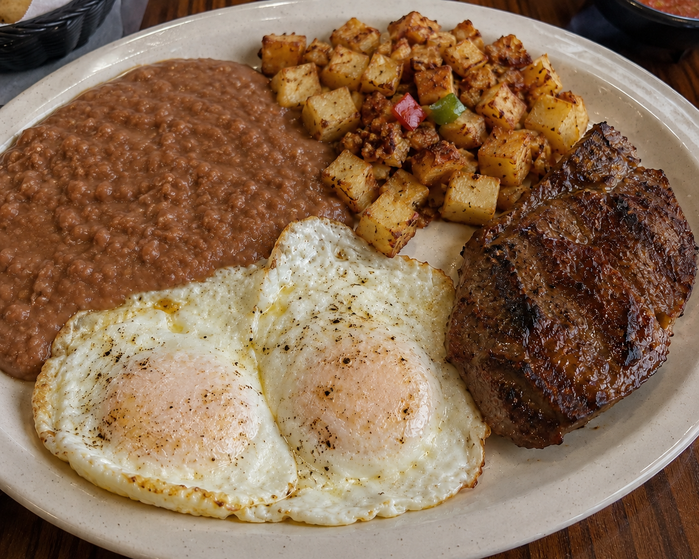 Steak and Eggs served on a white plate with a side of refried beans and potatoes